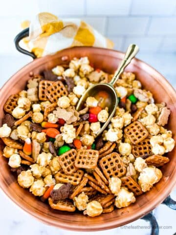 a large copper bowl of M and M halloween snack mix with a gold scoop inside and a gold and white napkin in the background