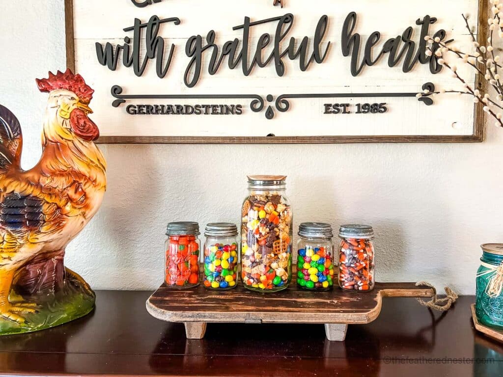 4 small and one large canning jars of fall and halloween candies and mixes on a wooden board on the top of a sideboard