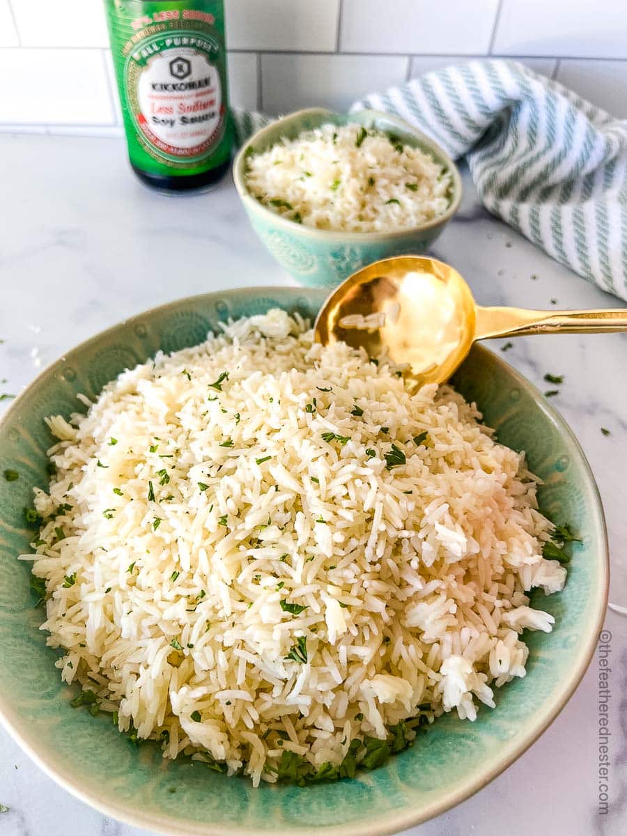 a large green bowl of Instant Pot Jasmine Rice with a golden serving spoon and a small bowl of Jasmine rice, a striped cloth, and a bottle of soy sauce in the background.