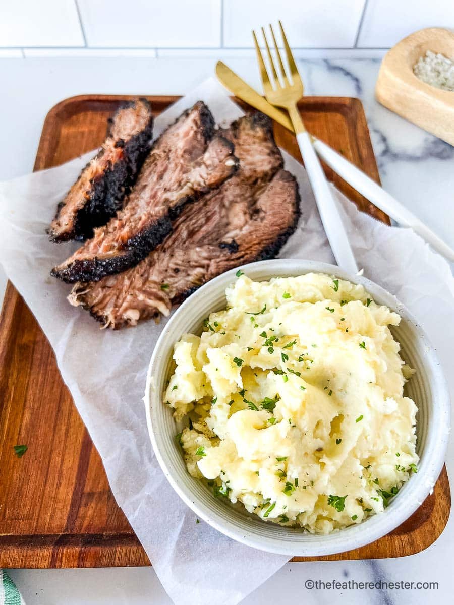 a white bowl of red mashed potatoes with slices of brisket on a wooden serving board with a striped napkin and gold fork and knife in the background.