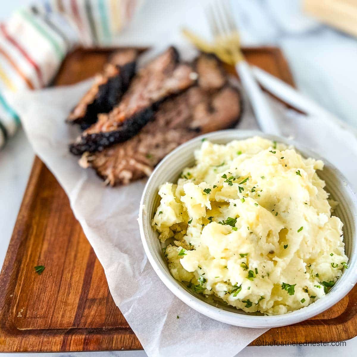 a white bowl of red mashed potatoes with slices of brisket on a wooden serving board with a striped napkin and gold fork and knife in the background.