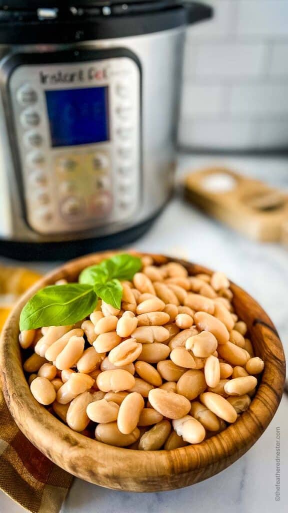 a wooden bowl of cooked cannellini beans with a sprig of basil on top and a gold and white napkin and instant pot in the background