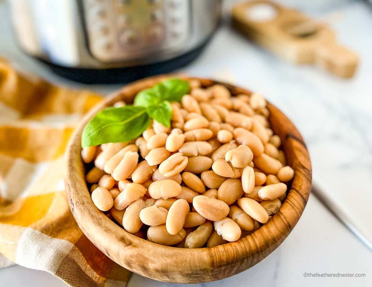 A wooden bowl of pressure cooked great northerni beans with a sprig of basil on top and a gold and white napkin and instant pot in the background.