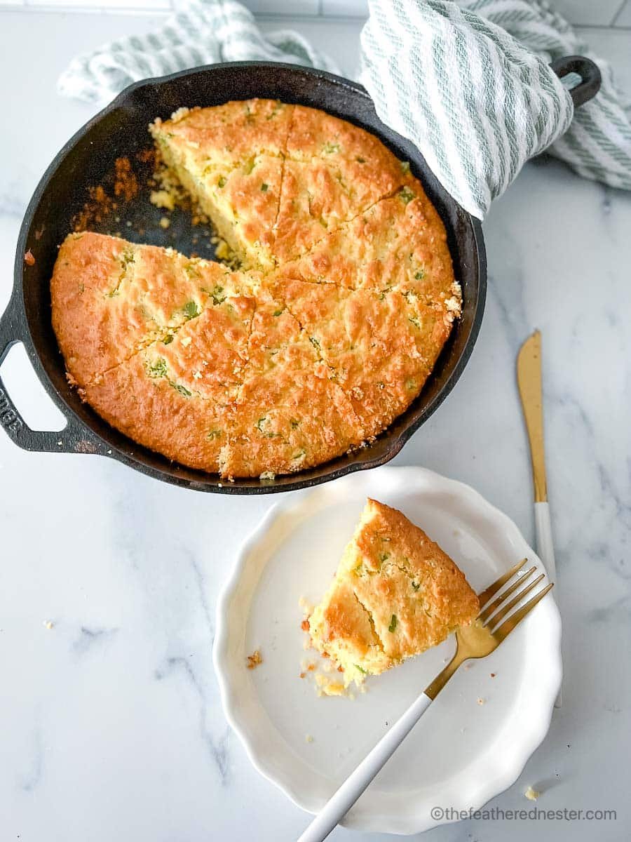 a white plate with a fork and a slice of jiffy jalapeno cheddar cornbread and a skillet with cornbread with striped cloth and bread knife in the background.