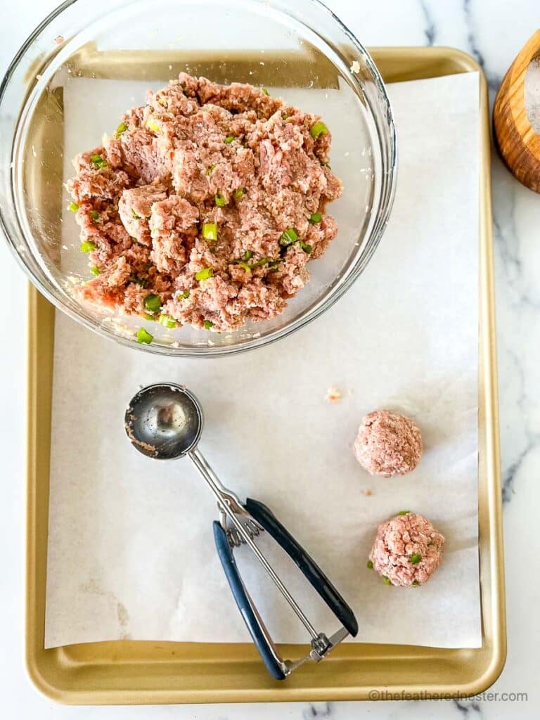 a baking sheet with parchment paper, a clear bowl of ground chicken meatballs, two rolled meatballs and a scoop.
