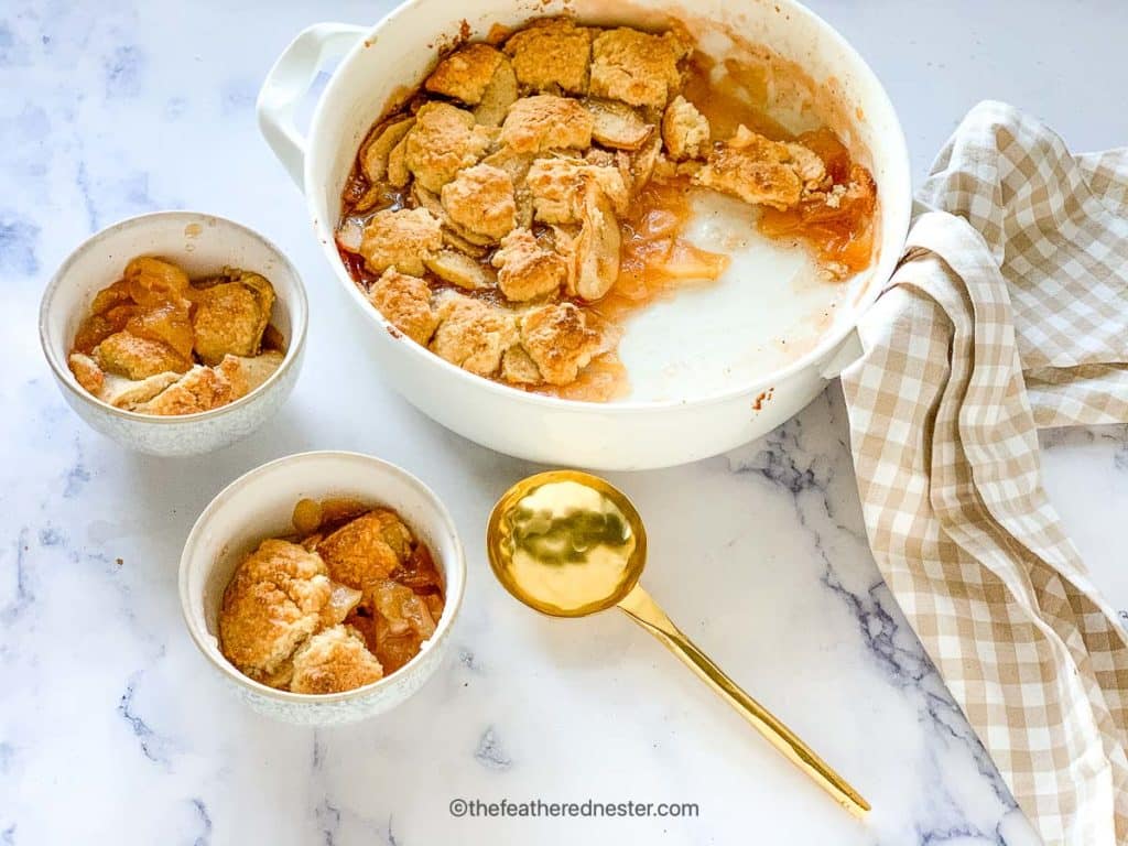 a white casserole dish of bisquick apple cobbler served into two white bowls with a gold serving spoon