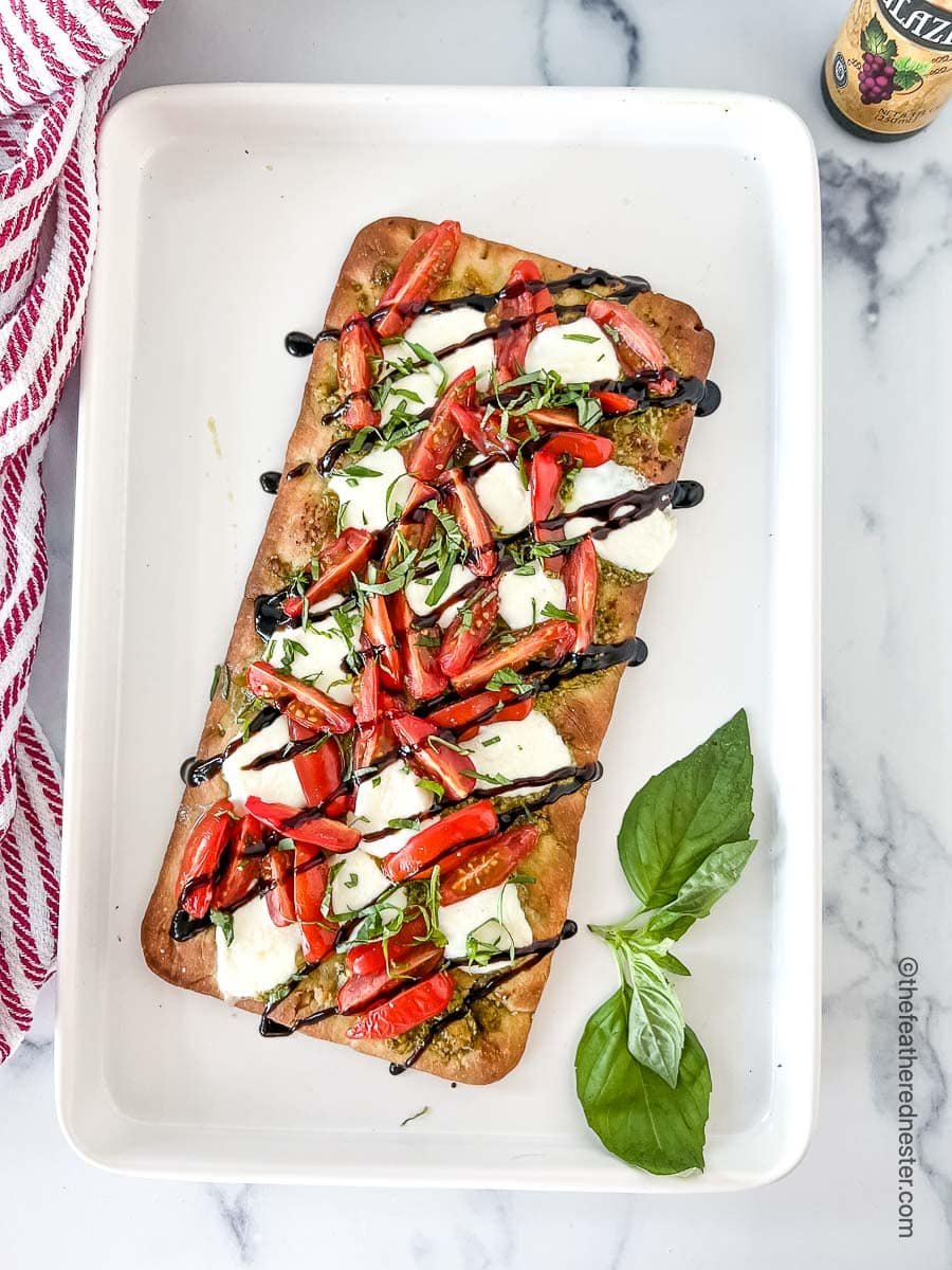 a white serving tray with a baked flatbread with mozzarella and tomato and sprig of fresh basil next to a red and white napkin.