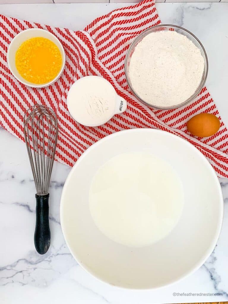ingredients for making Cornbread with self rising flour which contains melted butter, self rising flour, self rising cornmeal, egg, and whole milk with striped red kitchen towel, and a whisk at the side.