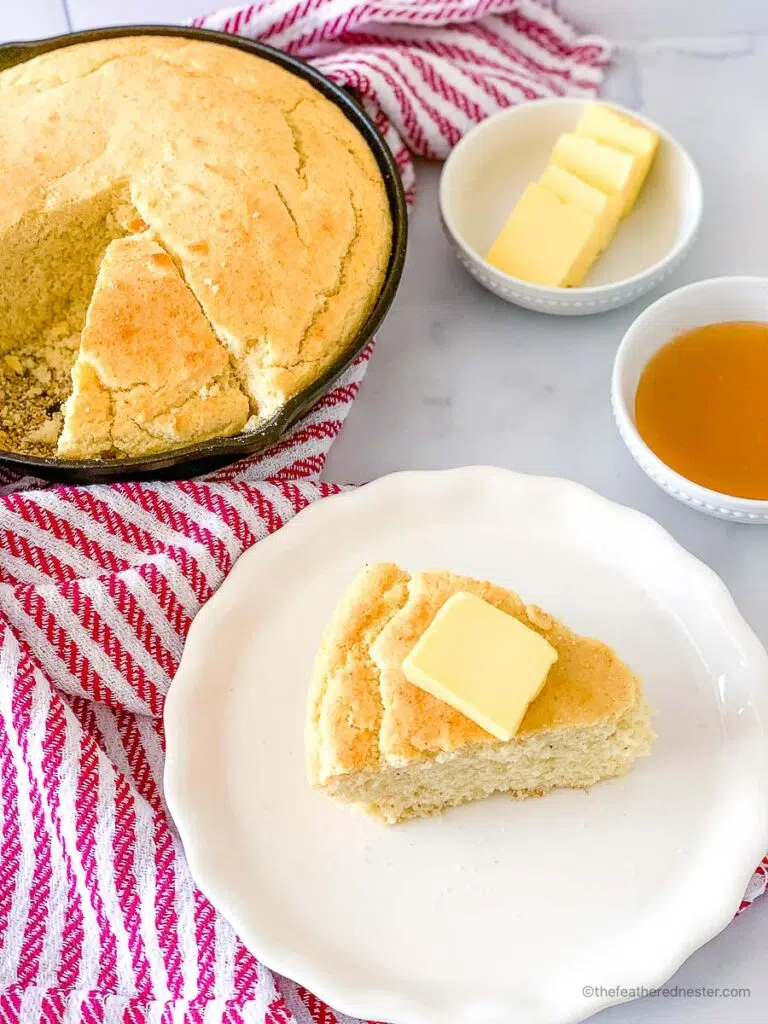 a white plate with a slice of cornbread with self rising flour with butter on top and a skillet with cornbread, striped red kitchen towel, bowls of sliced butter and honey at the back.