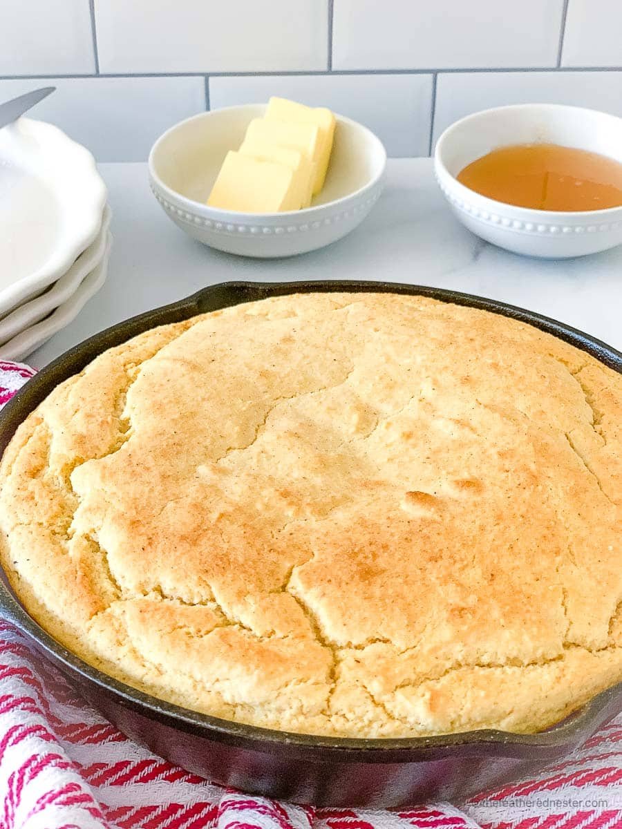 a cast iron skillet with Cornbread with self rising flour and a bowl of butter and honey, stack of plates, and striped red kitchen towel in the background.