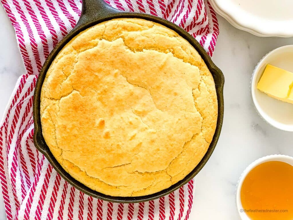 top view of cast iron skillet with cornbread with self rising flour and bowls of butter slices and honey, stack of plates, and a striped red cloth in the background.