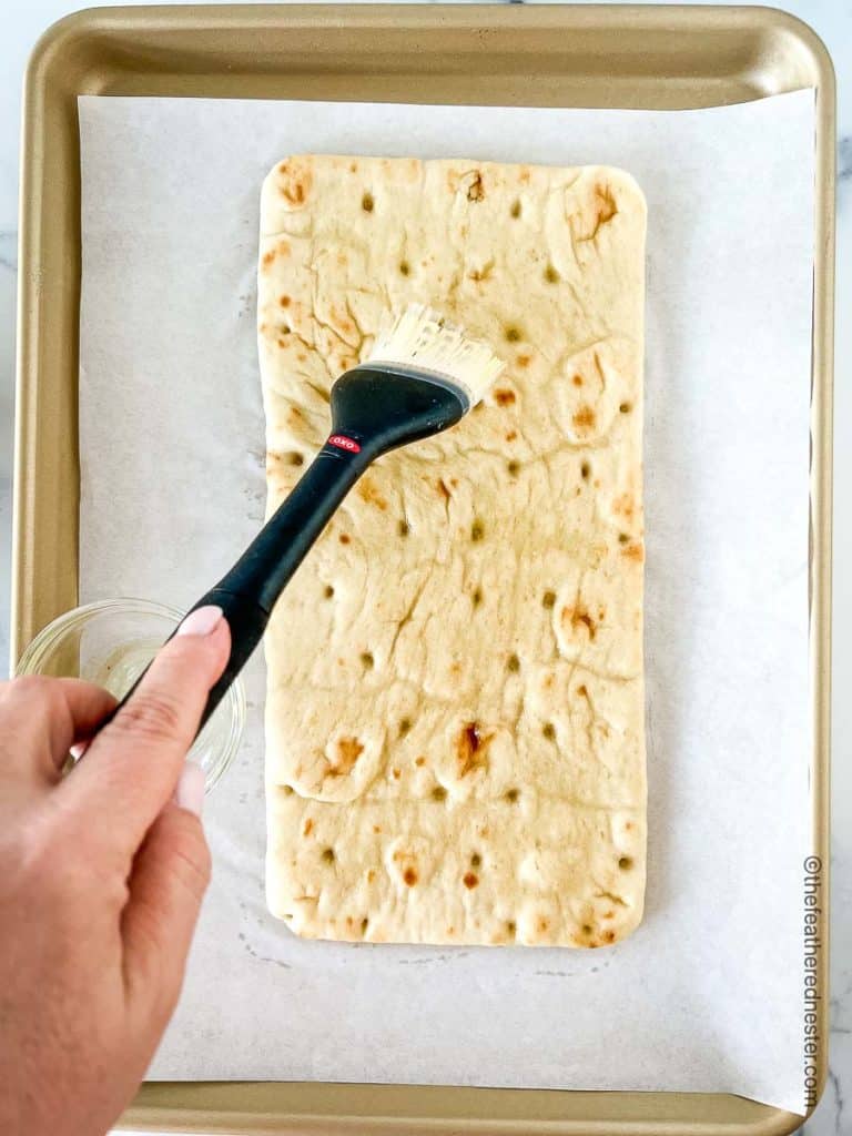 a hand brushing olive oil onto a flatbread on a parchment covered baking sheet