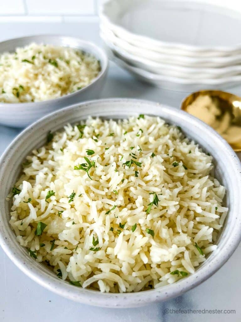 Two white bowls of Instant Pot Basmati Rice with a stack of white plates and golden serving spoon at the background.
