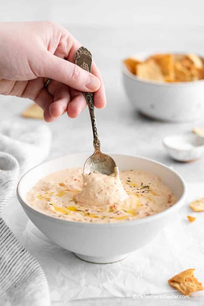 a hand holding a spoon scooping the roasted red pepper feta dip on a bowl with striped gray cloth, bowl of pita chips and salt in the background