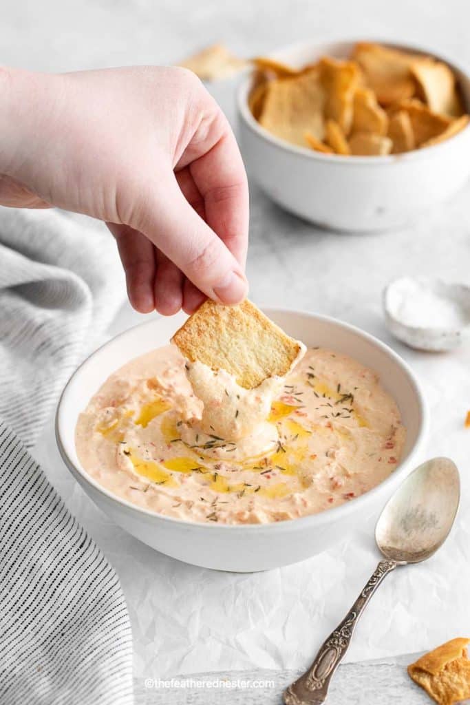 A hand holding a pita chips, dipping them into the Mediterranean-inspired roasted red pepper feta dip. A bowl of pita chips and salt are in the background