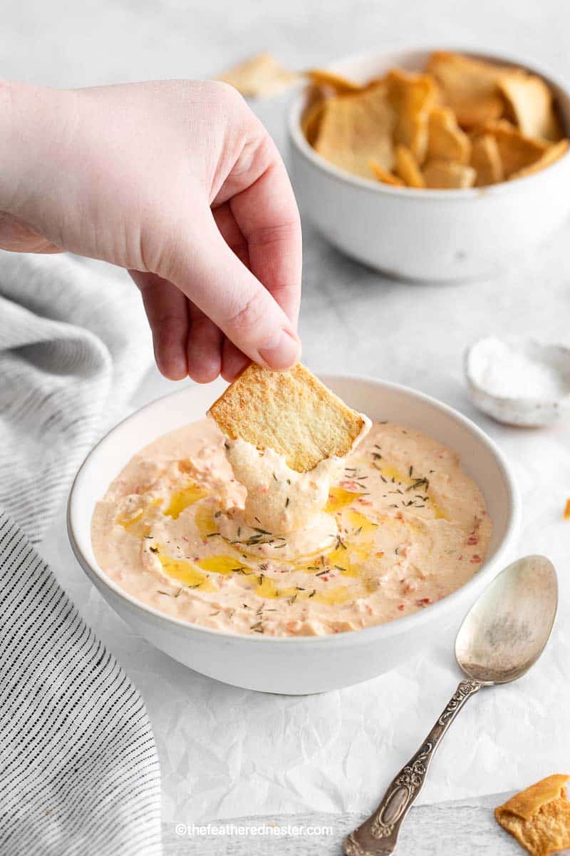a hand holding a pita chips dipping it to the Mediterranean-inspired roasted red pepper feta dip on a bowl with a spoon beside it and grapy striped cloth, and a bowl of pita chips and salt in the background