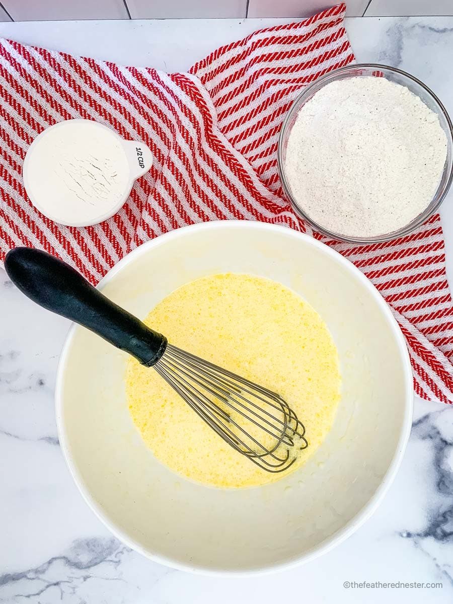 wet ingredients mixed using a whisk in a large mixing bowl and a cup of self rising flour, a bowl of self rising cornmeal and a striped red kitchen under them.