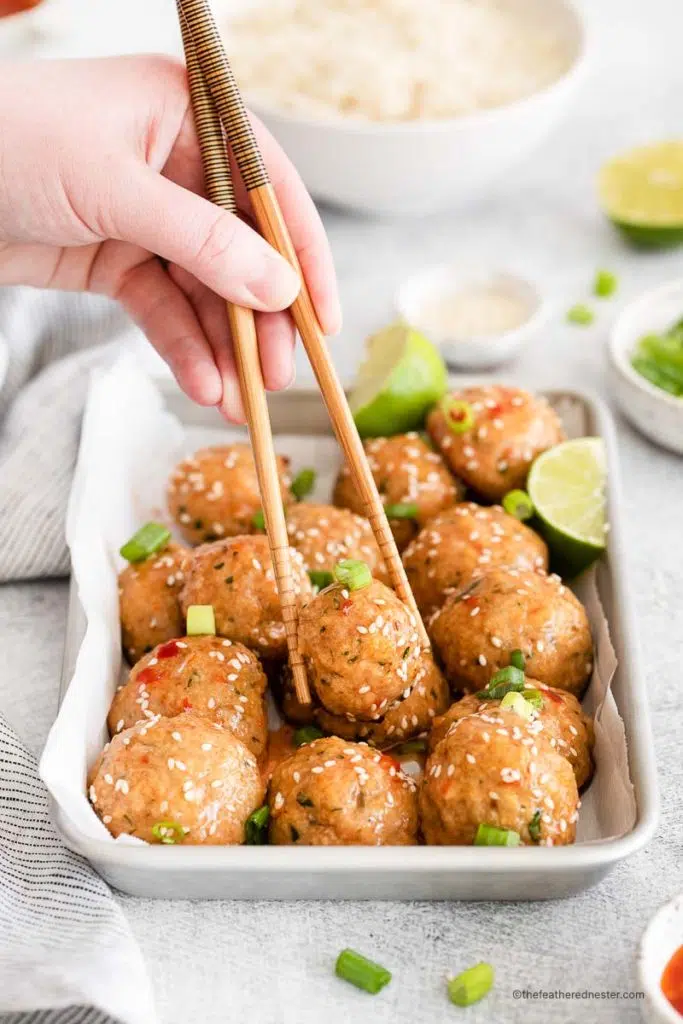 a hand holding chopsticks picking a meatballs placed in a baking sheet with dipping sauce and slice of lime and bowl of sesame seeds, bowl of green onions, and a bowl of rice in the background