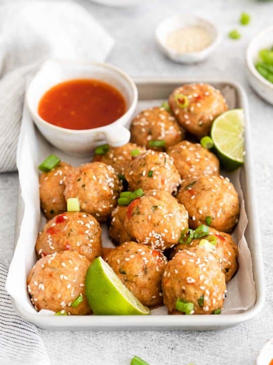 a baking sheet with sweet chili meatballs garnished with cilantro and sesame seeds and bowl of chili sauce and a slice of lime on its side with a gray cloth, bowl of green onions slices in the background