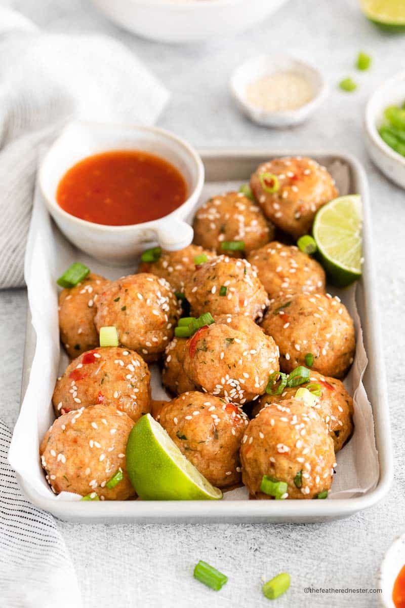 a baking sheet with sweet chili meatballs garnished with cilantro and sesame seeds and bowl of chili sauce and a slice of lime on its side with a gray cloth, bowl of green onions slices in the background.