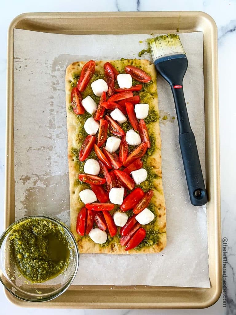 a baking sheet with a bowl of pesto sauce and a flatbread with pesto sauce, tomatoes, and mozzarella