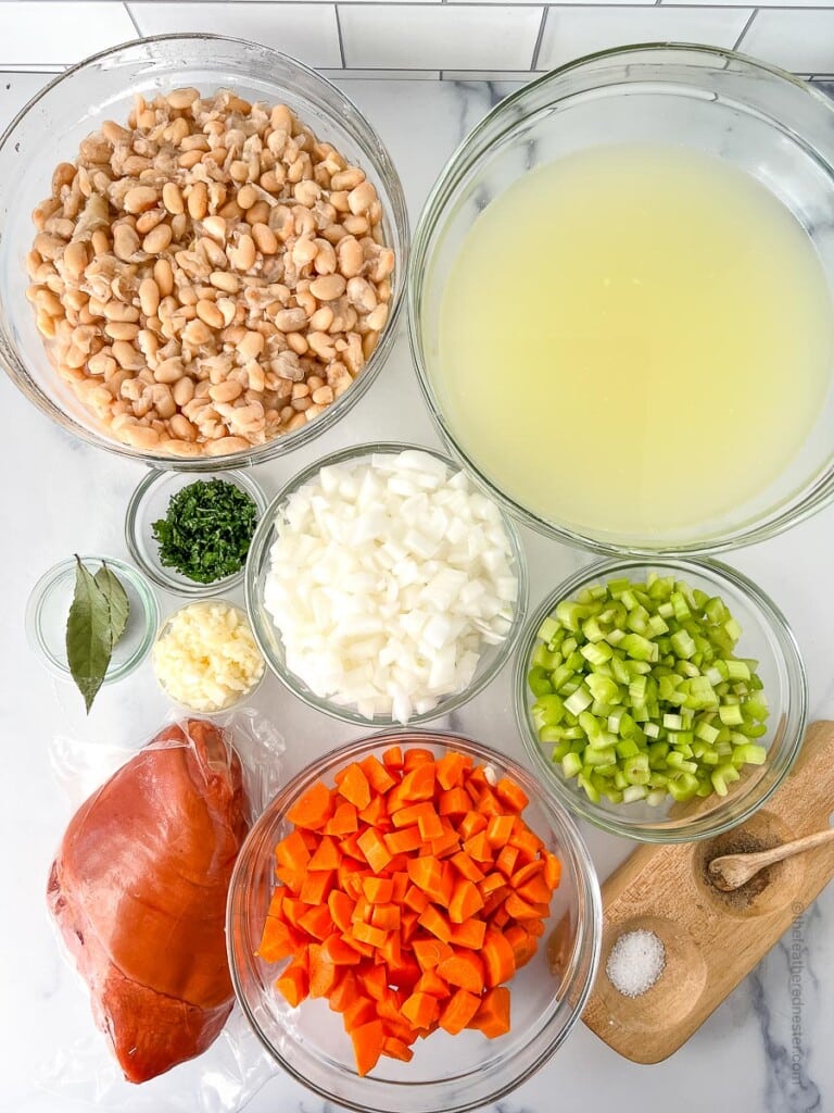 ingredients for making great northern bean soup which consists of pre soaked great norther beans, chicken broth, smoked ham hock, chopped onions, diced carrots, diced celery, dried oregano, minced garlic, bay leaves, salt and pepper.