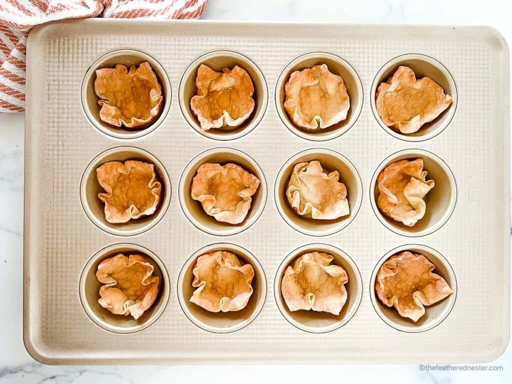 a muffin tin with baked wonton wrapper cups with a striped red kitchen towel on the side.