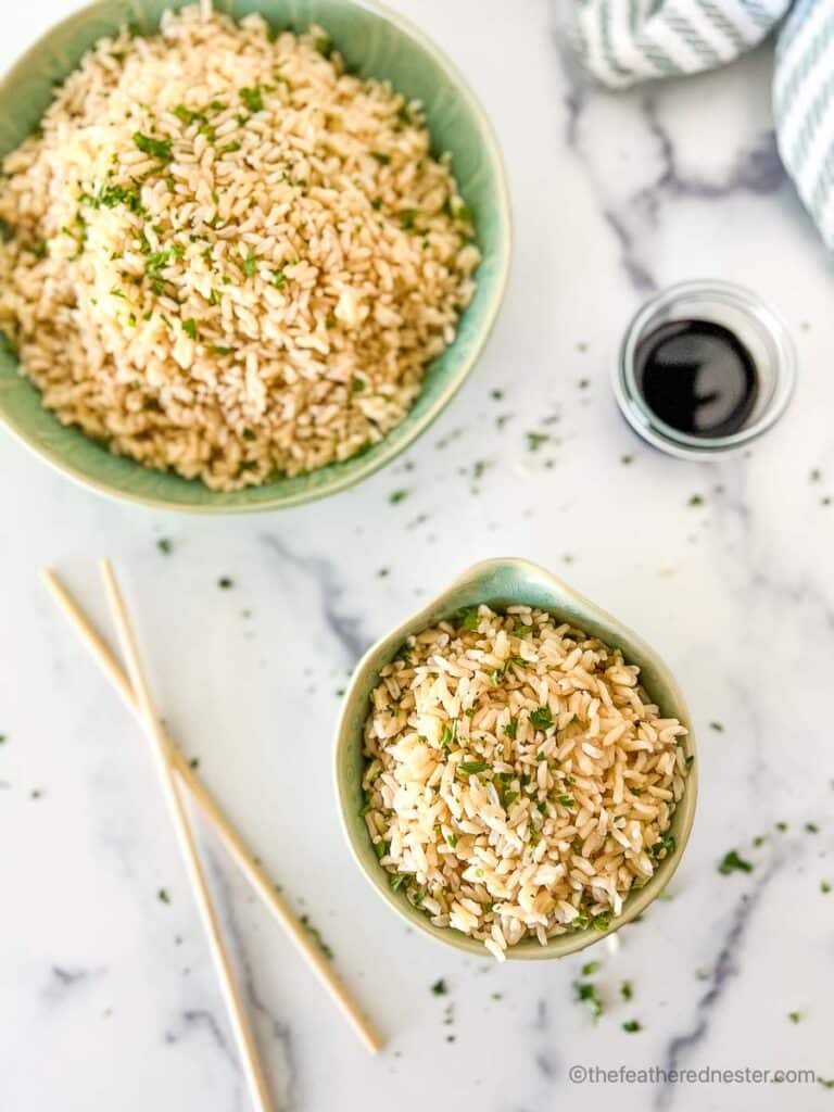 a small and large bowl of Instant Pot Brown Rice with chopped cilantros, chopsticks, soy sauce, and striped cloth in the background.