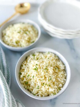 a white bowl of Instant Pot Coconut Rice and a striped cloth, another bowl of rice, golden serving spoon, and a stack of plates in the the background.