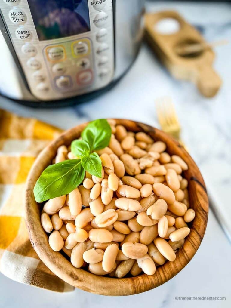 a wooden bowl of pressure cooked great northerni beans with a sprig of basil on top and a gold and white napkin and instant pot in the background