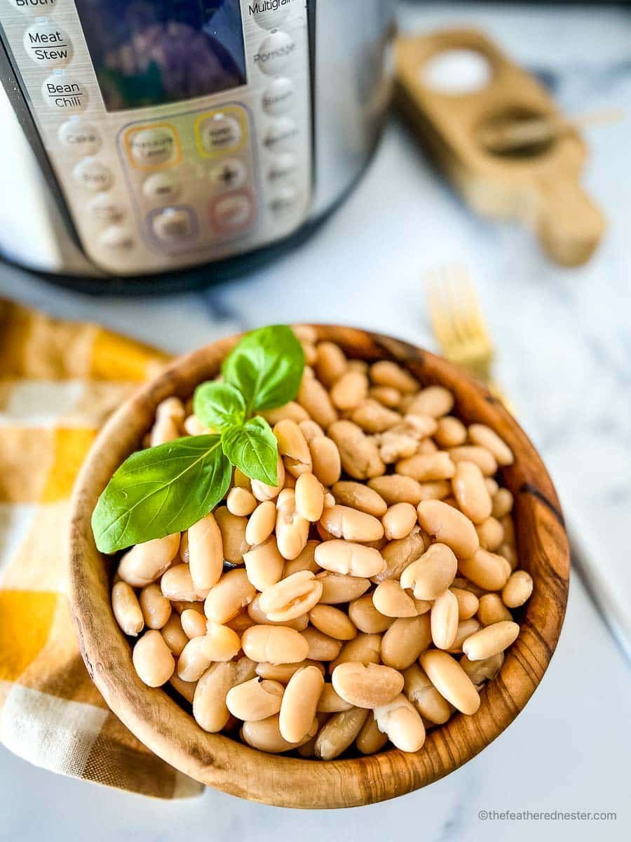 A wooden bowl of pressure cooked great northern beans with a sprig of basil on top and a gold and white napkin and instant pot in the background.