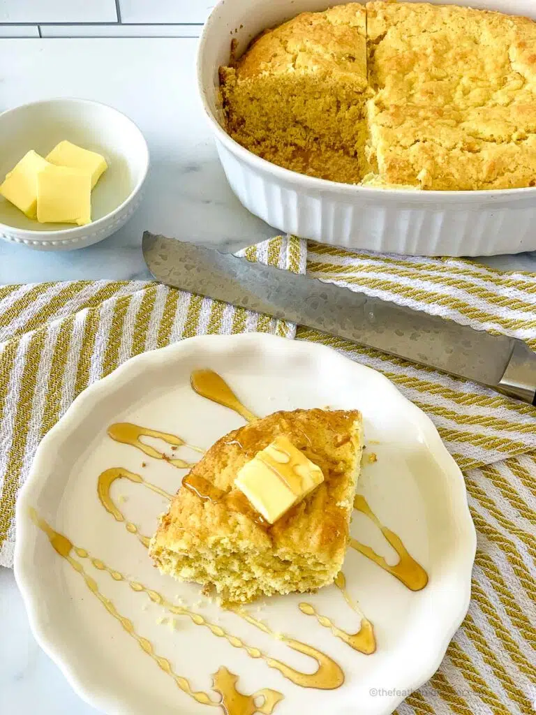 A white plate with a slice of Jiffy cornbread with creamed corn with butter on top, and a casserole with cornbread, knife, and a small bowl with butter in the back.