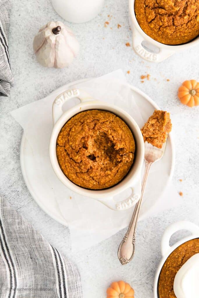a bowl of ramekin with pumpkin baked oats and a fork at the side placed on top of a white plate with another bowls of ramekins at the side.