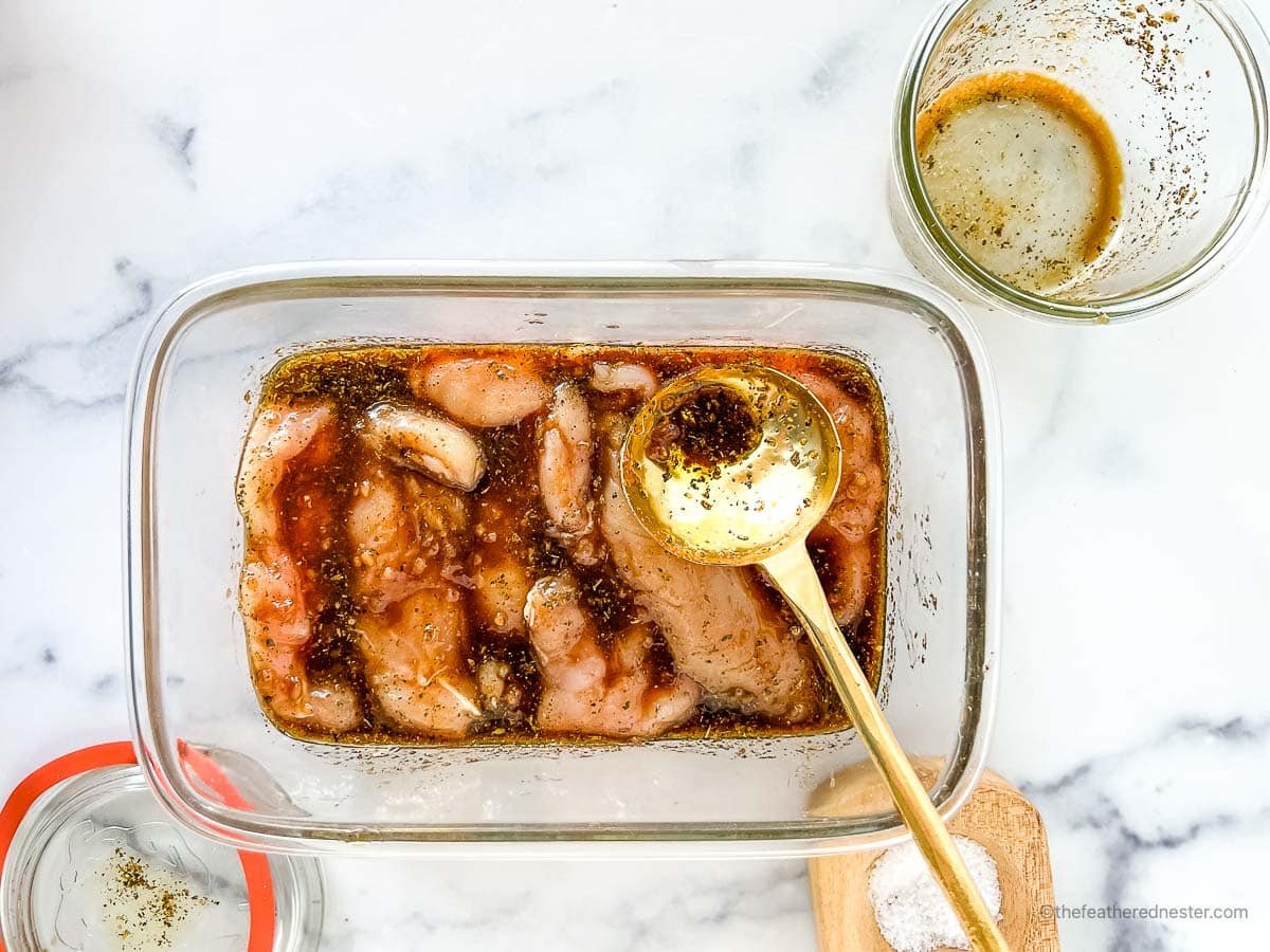 Marinated chicken placed in a glass container with a golden spoon on it and small bowls and salt cellar in the background.