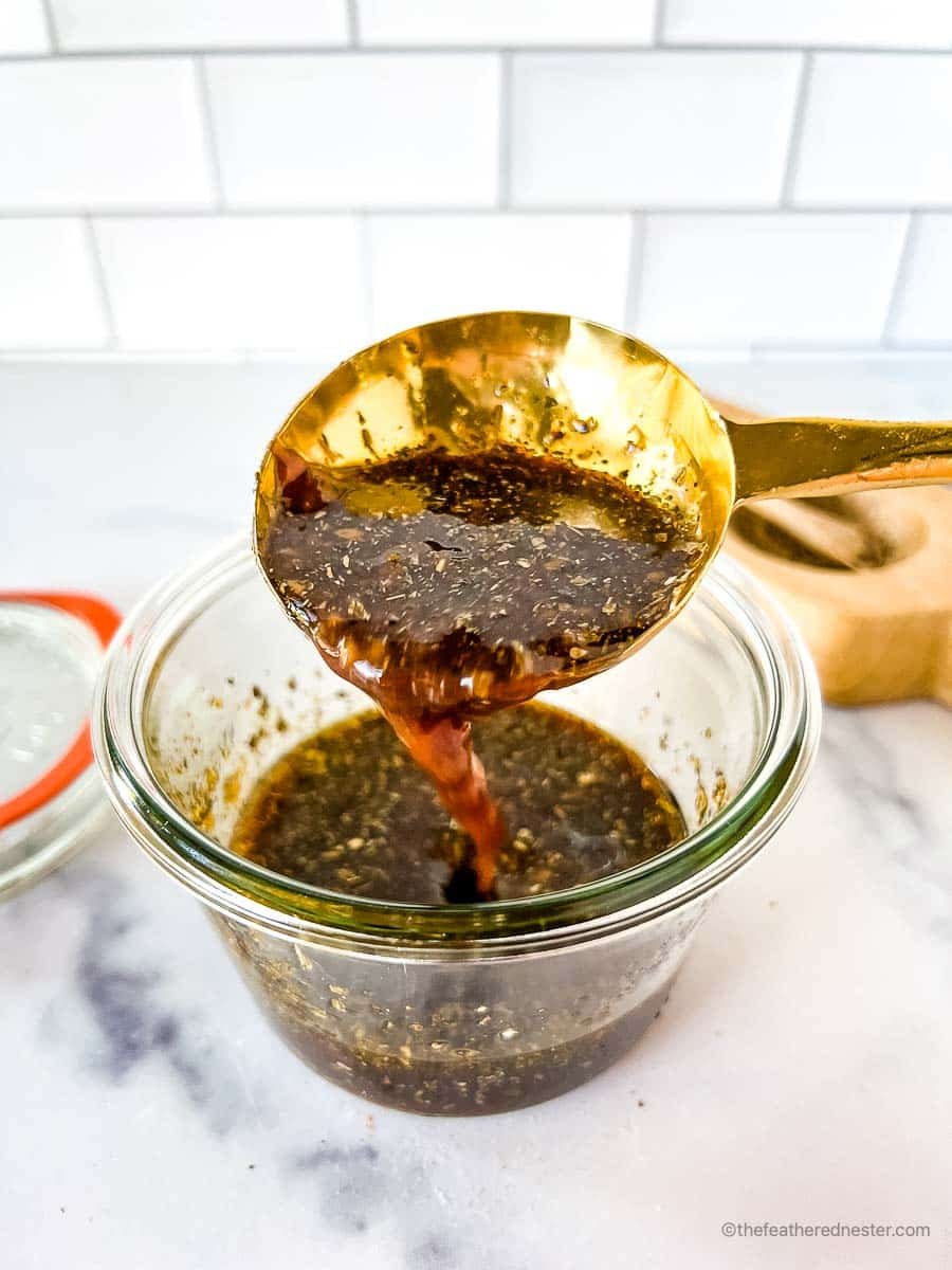 A golden spoon scooping the balsamic marinade for chicken in a glass jar with its lid and a salt cellar in the background.