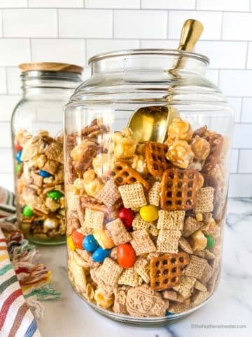 two jars of Chex mix with M&Ms, the other one has a golden scoop, and a striped kitchen towel in the background.