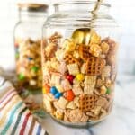 two jars of Chex mix with M&Ms, the other one has a golden scoop, and a striped kitchen towel in the background.