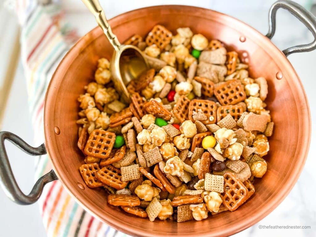 a wooden bowl full of Chex Mix with M&Ms with a golden scoop in it