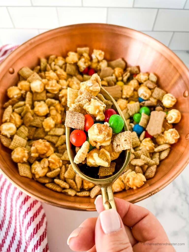 a hand holding a scoop of Chex mix with M&Ms and a wooden bowl full of it at the bottom.