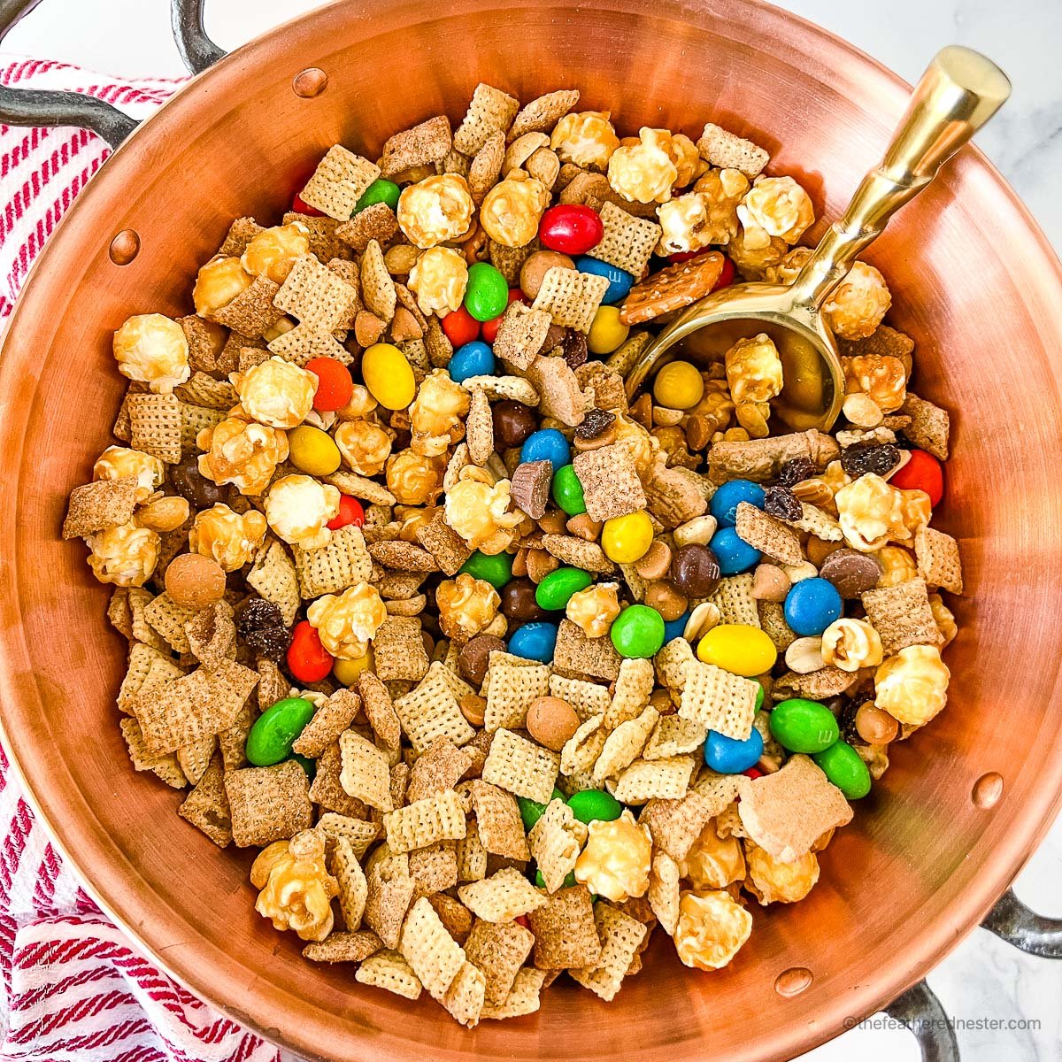 a close up photo of copper bowl with handle full of Chex Mix with M&Ms with a golden scoop on it, and a striped kitchen towel in the background.