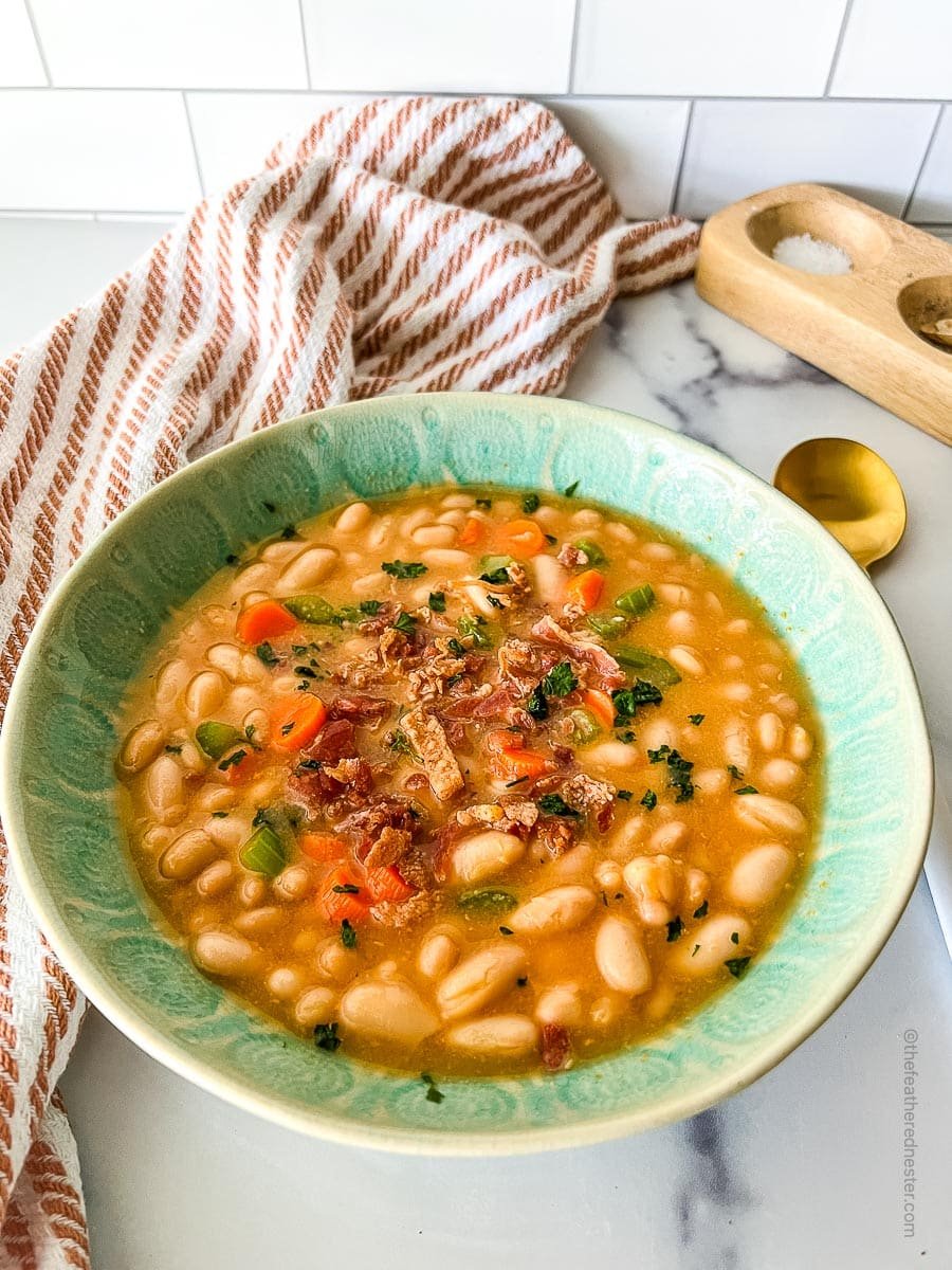 A green bowl with great northern bean soup and red kitchen towel, salt and pepper cellar, and golden spoon at the back.