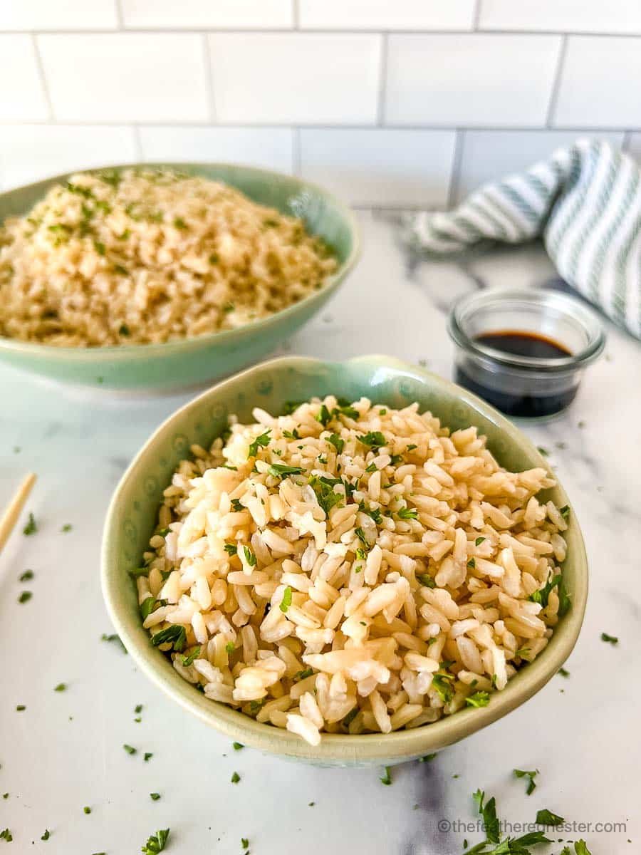 A small and large green bowls with brown rice garnished with chopped cilantro with a soy sauce in a small bowl, striped cloth in the background.