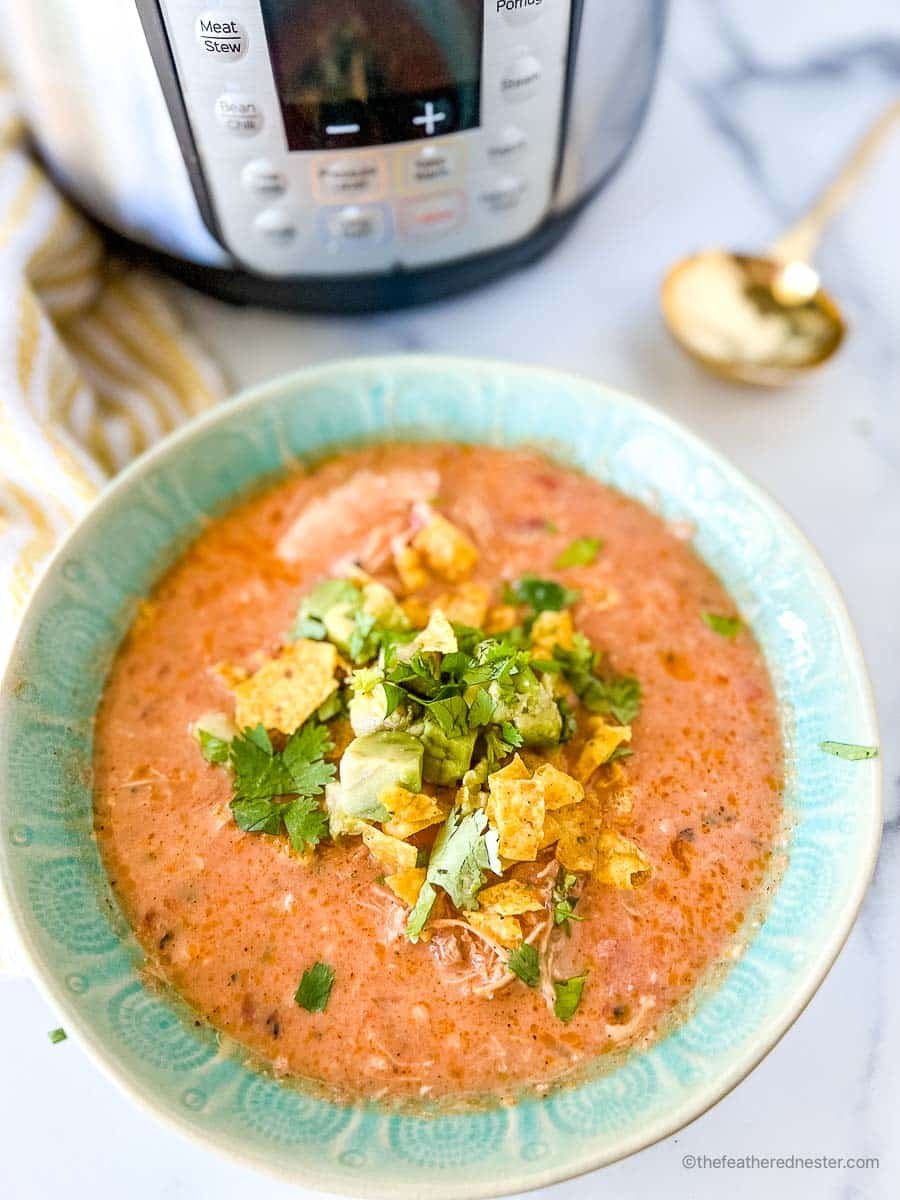 A slightly close up photo of a green bowl of instant pot creamy chicken tortilla soup with an instant pot, white and yellow napkin, and gold spoon.