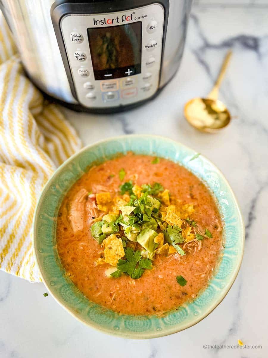 a green bowl of instant pot creamy chicken tortilla soup with an instant pot, white and yellow napkin, and gold spoon.