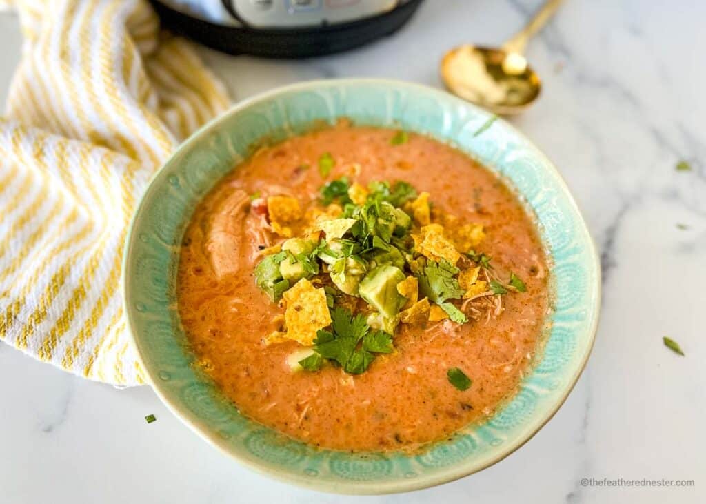 a horizontal photo of a green bowl with Instant Pot Chicken Tortilla Soup with a striped yellow cloth, Instant Pot, and a golden spoon at the side.