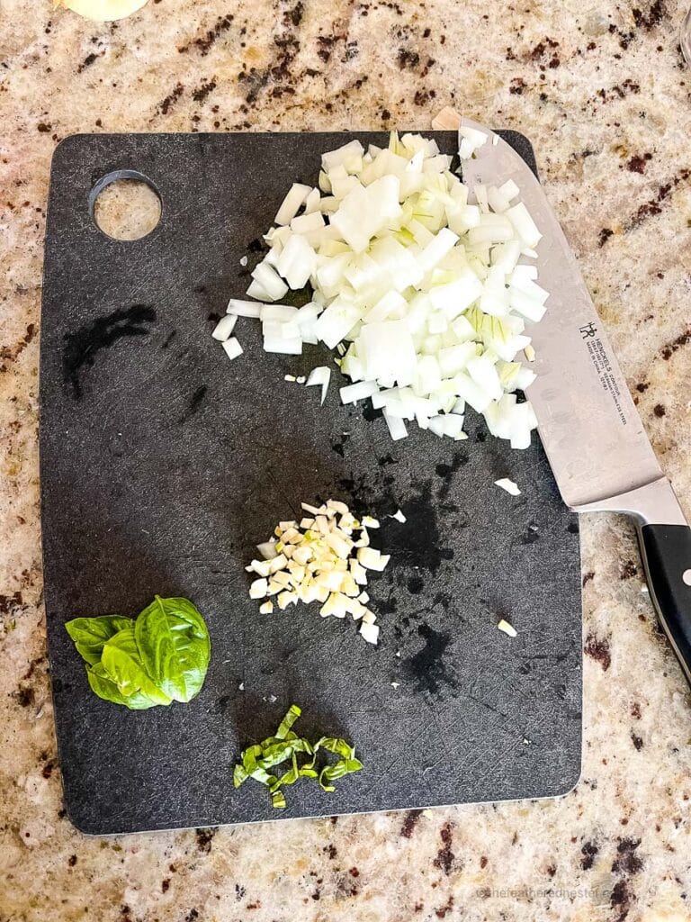 chopped onions, garlic and basil leaf on top of a black chopping board with a knife.
