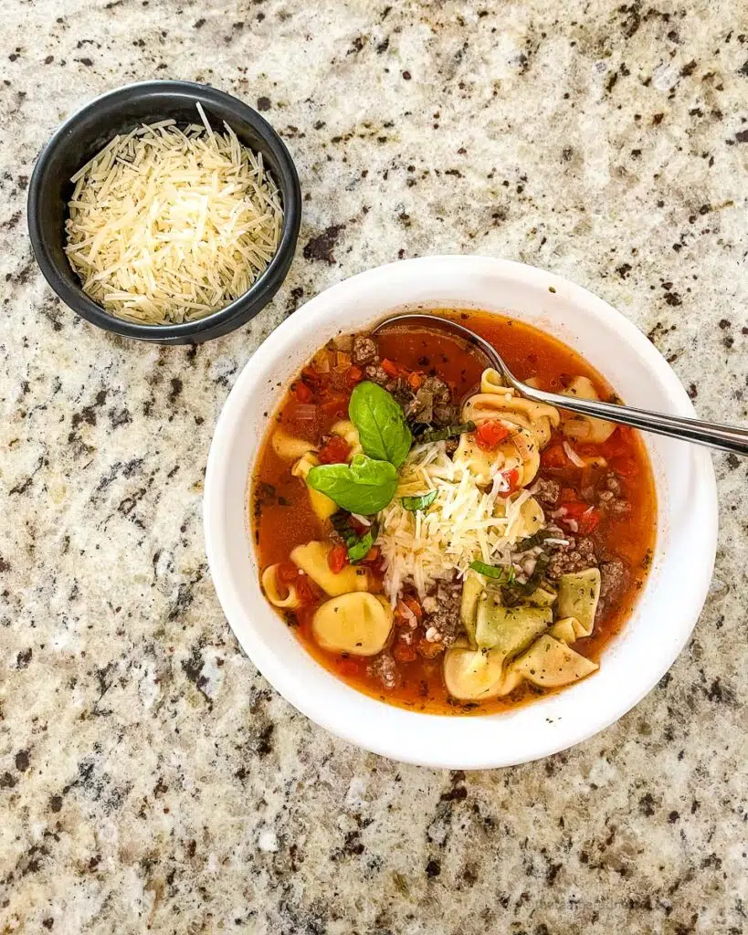 a bowl of Instant Pot Tortellini Soup with a little bowl of shredded cheese at the side.