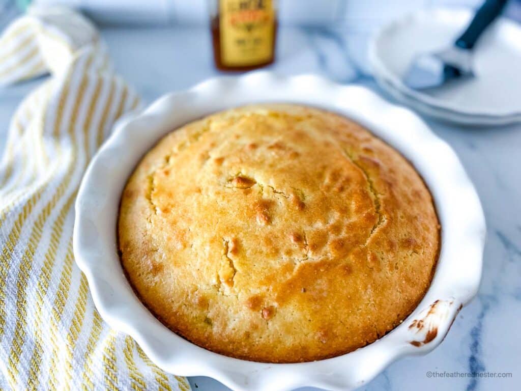 a casserole dish of Jiffy cornbread with buttermilk with a striped yellow cloth, bottle of honey, cake slicer, and a plate in the background.