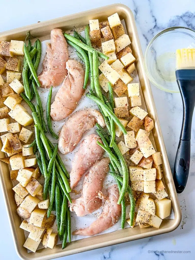 chicken tenders, asparagus, and potatoes, seasoned with all-purpose seasoning, all lined in a parchment paper on a baking sheet and a basting brush and oil at the side.