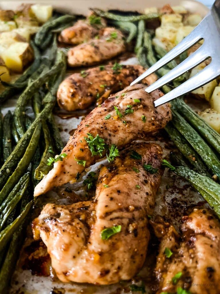Oven baked marinated chicken tenders with a fork poking it and baked asparagus and potatoes beside it.
